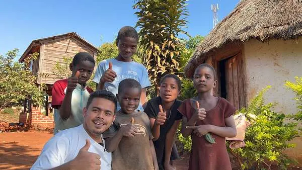 Saif with a group of children giving a thumbs up outside a thatched home
